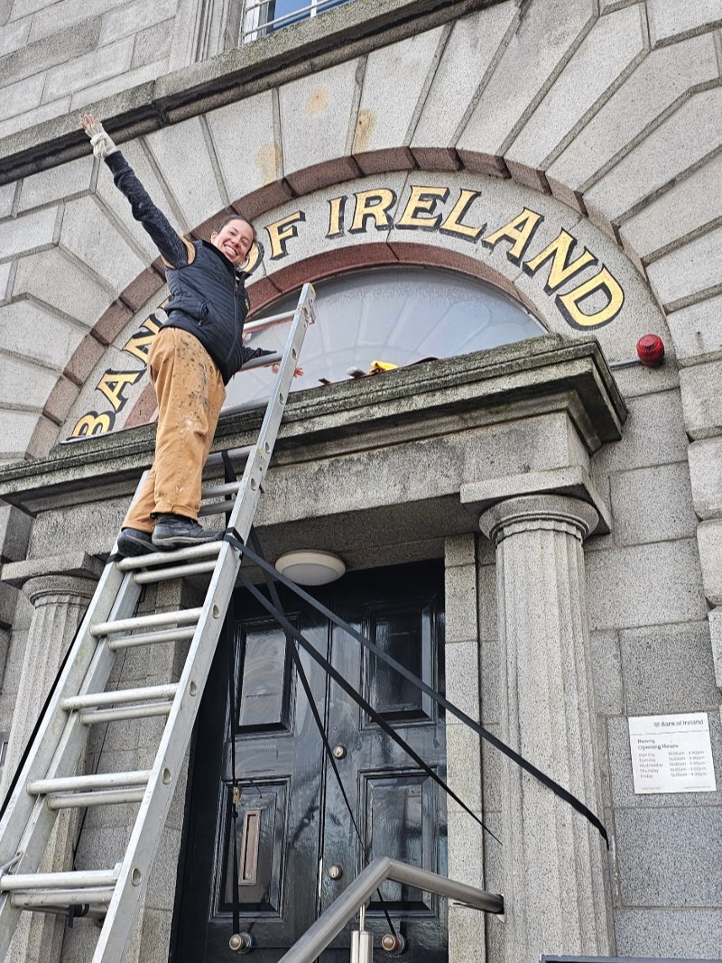 Joha Muñe standing in front of the gilded Bank of Ireland sign in Lurgan after completing gold leaf work.